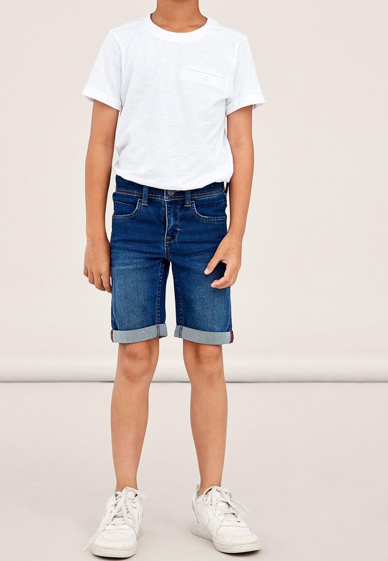 T-shirt blanc à manches courtes avec une poche poitrine, accompagné d'un short en denim bleu retroussé. Des baskets blanches complètent la tenue.