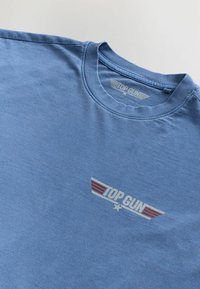 T-shirt in cotone azzurro chiaro con scollo a girocollo e logo stampato "TOP GUN" con accenti rossi a forma di ala sul lato sinistro del petto.