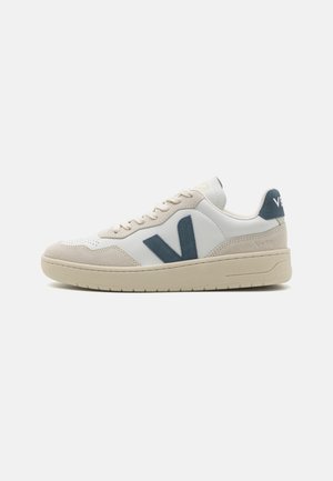 Baskets basses avec semelle beige, dessus en cuir blanc et en daim, et logo "V" gris foncé sur le côté.