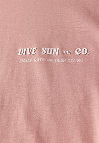 Světle růžová bavlněná látka s textem "DIVE SUN and CO." v tučných bílých písmenech, pod nímž se nachází menším bílým písmem "DAILY RAYS and DEEP DIVING."