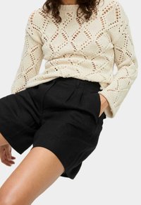 Pull crème en tricot avec un motif en treillis ouvert, associé à un short noir taille haute avec des plis et des poches latérales.