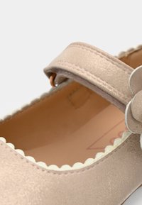 Gros plan d'une chaussure Mary Jane beige avec bord festonné, bride ajustable et fleur décorative sur le côté.