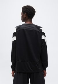 Άτομο με κοντά σγουρά μαλλιά που φοράει μαύρο πουλόβερ με μακριά μανίκια και λευκές ρίγες στους ώμους, εμφανιζόμενο από πίσω.