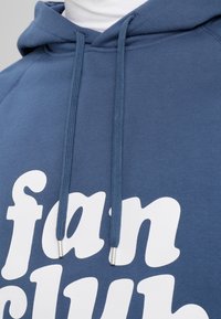Blå hoodie med en vit "fan club"-grafik. Har dragsnören med metallspetsar och ribbade muddar. Mjuk, texturerad tyg.