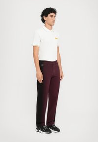 T-shirt polo blanche avec des accents jaunes, associée à un pantalon bordeaux foncé. Le modèle porte des baskets noires, se tenant devant un fond blanc uni.