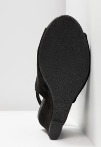 Semelle en suède noir avec grip en caoutchouc texturé, présentée debout contre un mur et un sol blancs.