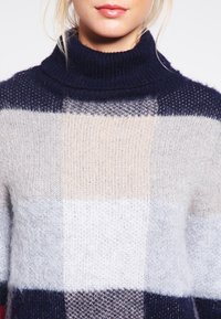 Kvinna iklädd en mjuk, lurvig stickad polotröja med ett stort rutmönster i marinblått, beige, ljusgrått och vitt.