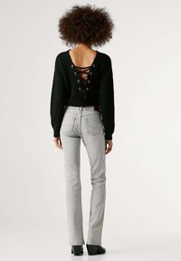 Μαύρο πλεκτό πουλόβερ με βαθύ V στην πλάτη, σχέδιο με κορδόνια και μακριά μανίκια, συνδυασμένο με στενά ανοιχτόγκρι τζιν και μαύρα μπότες με τακούνι.