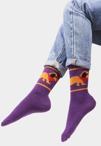 Fioletowe skarpetki z pomarańczowym wzorem psa w czerwonej bandanie, z prążkowanymi mankietami i kontrastującymi kolorami w zestawieniu z dżinsami.