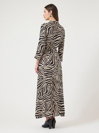 Γυναίκα με μακρύ φόρεμα σε μαύρο και μπεζ με ζεβρέ σχέδιο, με μανίκια μέχρι τους τρεις τέταρτους, που στέκεται σε μαύρα παπούτσια με τακούνι, κοιτώντας προς τα πλάγια.