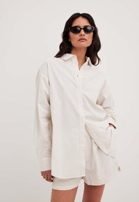 White striped shirt with a collar, long sleeves, and buttons, paired with matching shorts. Model wears black sunglasses. Smooth texture.