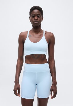 Femme aux cheveux courts debout, tournée vers l'avant, portant un brassière de sport bleu clair et un short de vélo taille haute assorti sur un fond uni.