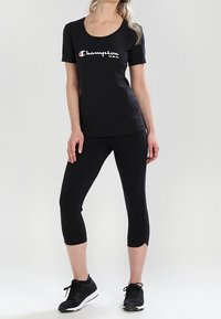 Svart bomullströja med rundad hals och vit "Champion U.S.A."-logga, i kombination med svarta korta leggings och svarta träningsskor.