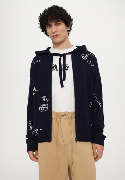 Gilet à capuche marine avec texte et motifs brodés en blanc, associé à un t-shirt blanc et un pantalon beigne à cordon. Silhouette ajustée.