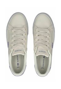 Trampki Lacoste mają kremową, syntetyczną cholewkę, fioletowy napis marki, płaskie sznurówki oraz teksturowaną gumową podeszwę. Okrągły kształt noska.