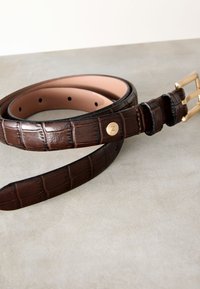 Brunt krokodilläderbälte med slät textur, spänne i guldfärgat metall och fem justeringshål, som visar en polerad yta.