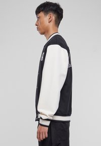 Veste varsity noire et blanche avec une fermeture à boutons, des manches contrastées, des poignets côtelés et un logo brodé sur la poitrine.