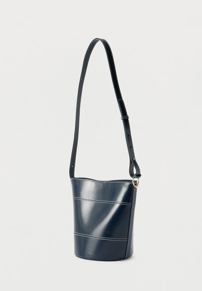 Τσάντα κουβάς από δέρμα σε ναυτικό μπλε χρώμα με λευκές ραφές και ρυθμιζόμενο λουρί ώμου, κρεμασμένη σε απλό φωτεινό φόντο.