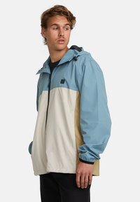 Men's color-blocked jacket featuring a light blue upper, cream lower section, and beige sides. Made of lightweight, water-resistant material.