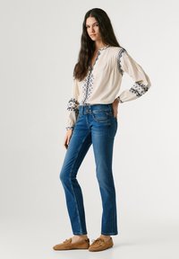 Woman with long dark hair wearing a cream blouse with blue embroidery, blue jeans, and brown loafers, standing with one hand on her hip.
