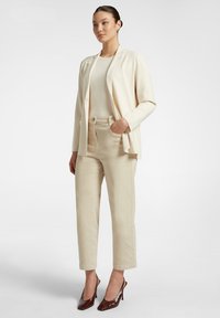 Blazer beige chiaro sopra un top abbinato e pantaloni high-waisted di colore chiaro. Tacchi marroni completano l'outfit. Tessuto dalla texture liscia.