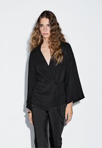 Žena s dlouhými vlnitými vlasy, oblečená v černém topu ve stylu wrap s širokými rukávy a odpovídajících černých kalhotách na jednobarevném bílém pozadí.
