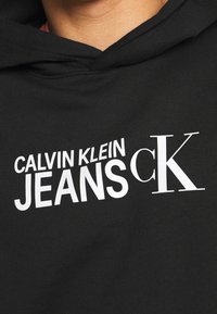 Felpa nera con la scritta "CALVIN KLEIN JEANS" in bianco, caratterizzata da un grande logo stampato e un design classico a pullover. Tessuto morbido.