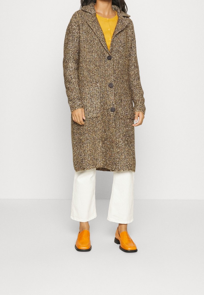 Dlouhý hnědý a krémově texturovaný svetr na knoflíky, kombinovaný se žlutým topem a bílými kalhotami, doplněný jasně oranžovými botami.