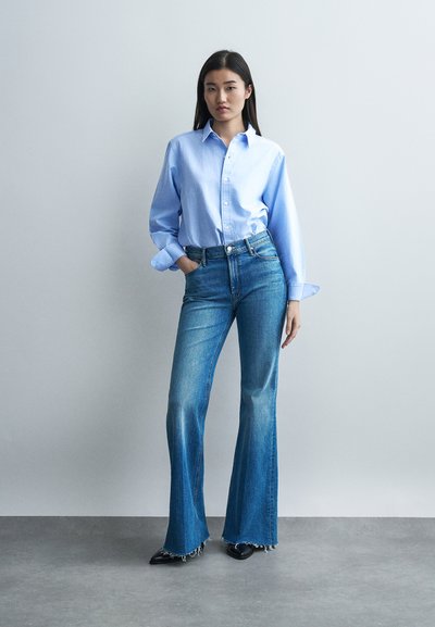 Γυναίκα που στέκεται με το ένα χέρι στην τσέπη, φοράει ανοιχτό μπλε πουκάμισο με κουμπιά και ψηλόμεσα τζιν καμπάνα με ξεφτισμένα τελειώματα σε γκρι φόντο.