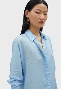 Camicia azzurra chiara con vestibilità rilassata, caratterizzata da dettagli ricamati distribuiti in modo casual e un colletto classico. Tessuto morbido, maniche lunghe.