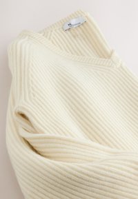 Krémový rebrovaný sveter s výstrihom do V a textúrovanou látkou, s viditeľnou etikou pri výstrihu. Jemný, teplý materiál.