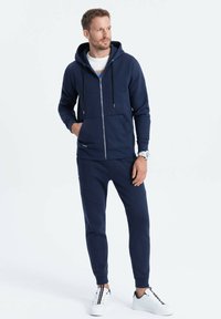 Marine blauwe zip-up hoodie met twee voorzakken en trekkoorden, gecombineerd met bijpassende tapered joggers. Gekleed met witte sneakers.