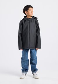 Black waterproof jacket with a hood, front zipper, and two side pockets, worn over light blue jeans and white sneakers.