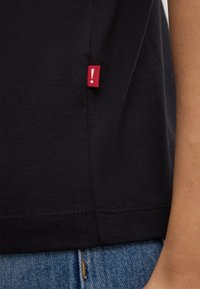 Schwarzes Baumwoll-T-Shirt mit abgerundetem Saum, versehen mit einem roten Etikett mit einem weißen Ausrufezeichen an der Seite. Denim-Jeans sind unten sichtbar.
