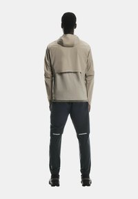 Gaišs beiges jaka ar kapuci un pagarinātu muguru, savienota ar tumšām biksēm ar atstarojošiem akcentiem. Gluda tekstūra un moderna dizaina izskats.