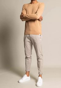 Beżowy sweter z dzianiny z długimi rękawami, zestawiony z jasnoszarymi zwężanymi spodniami i białymi trampkami. Gładka faktura, dopasowany fason, minimalistyczne oznaczenia.