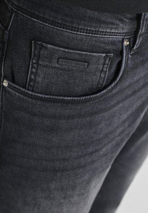 Nahaufnahme einer schwarzen Denim-Jeans vorne mit einer kleinen Armbanduhr und einem Etikett mit der Aufschrift Antony Morato.