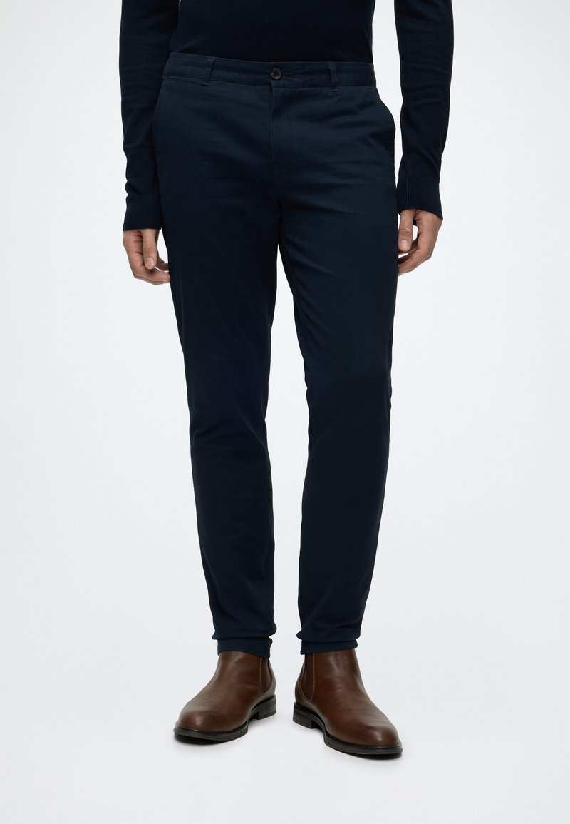 Homme portant un pantalon slim bleu marine et des bottines en cuir marron, debout sur fond blanc.