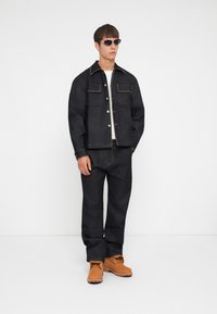 Donkerdenim jack met lichte stiksels, twee voorknosjes, gecombineerd met bijpassende donkerdenim broeken en tan werklaarzen. Zonnebril maakt de look compleet.