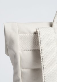 Beige tygväska med en strukturerad yta, utrustad med en bred rem och vävda detaljer, som visar en minimalistisk design och robust konstruktion.