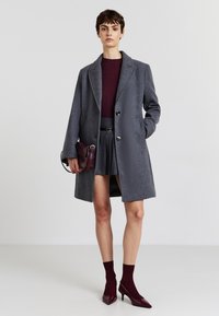 Cappotto di lana grigio con un design su misura, caratterizzato da due bottoni, abbinato a un top a coste bordeaux, una gonna plissettata e accessori coordinati.