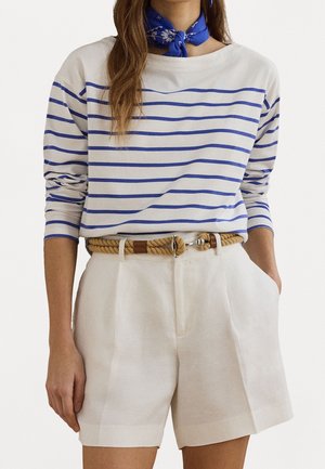 Frau trägt ein blau-weiß gestreiftes Langarmshirt, beige hochtaillierte Shorts mit einem Seilgürtel und ein blau gemustertes Halstuch.