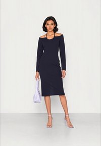 Vestido ajustado azul marinho com mangas longas, design de ombro à mostra, textura suave, comprimento até aos joelhos, combinado com saltos lilás claros e uma mala.