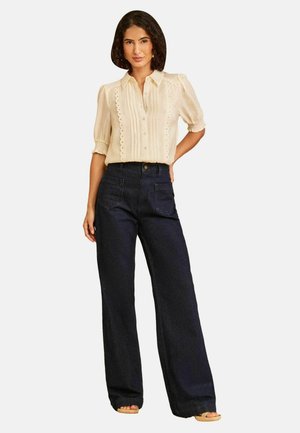 Kvinde iført en cremefarvet kortærmet bluse med blonde detaljer og højtaljede, vidde, mørke jeans, stående med den ene hånd på hoften.