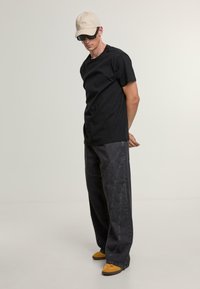 Black cotton t-shirt, loose gray denim trousers with curved stitching, beige cap, and yellow shoes, standing on a neutral background.