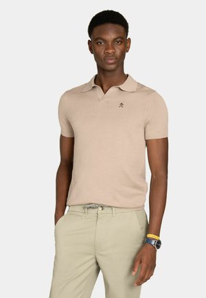 Hombre con camisa polo beige de manga corta y pantalones caqui claros, de pie con una mano en el bolsillo contra un fondo liso.