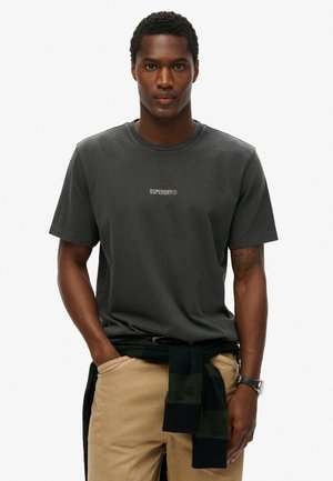 Mørkegrå bomulds-t-shirt med rund halsudskæring og lille hvid "Superdry" logo. Bæres med beige bukser og en grønternet sweater bundet om livet.