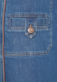 Un bolsillo de mezclilla azul con costuras marrones, una solapa asegurada por un botón de metal marcado y un borde de cuero marrón a lo largo del borde izquierdo.