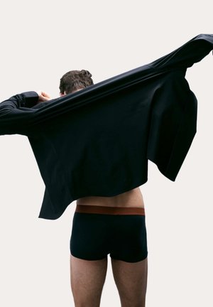 Una maglietta nera a maniche lunghe viene rimossa, rivelando pantaloni corti scuri con una vita marrone. Tessuto liscio con texture minimal, vista posteriore.