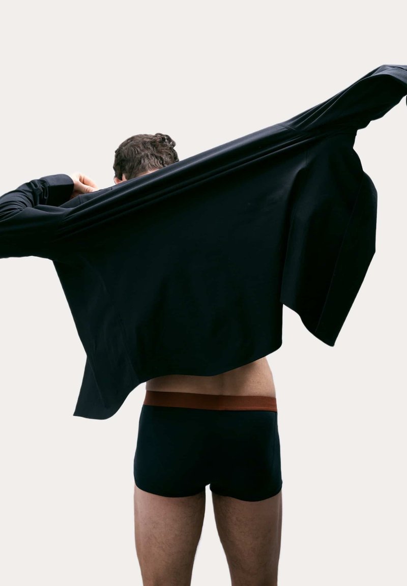 Camiseta negra de manga larga siendo retirada, revelando pantalones cortos oscuros con una cinturilla marrón. Tela suave con mínima textura, vista trasera.